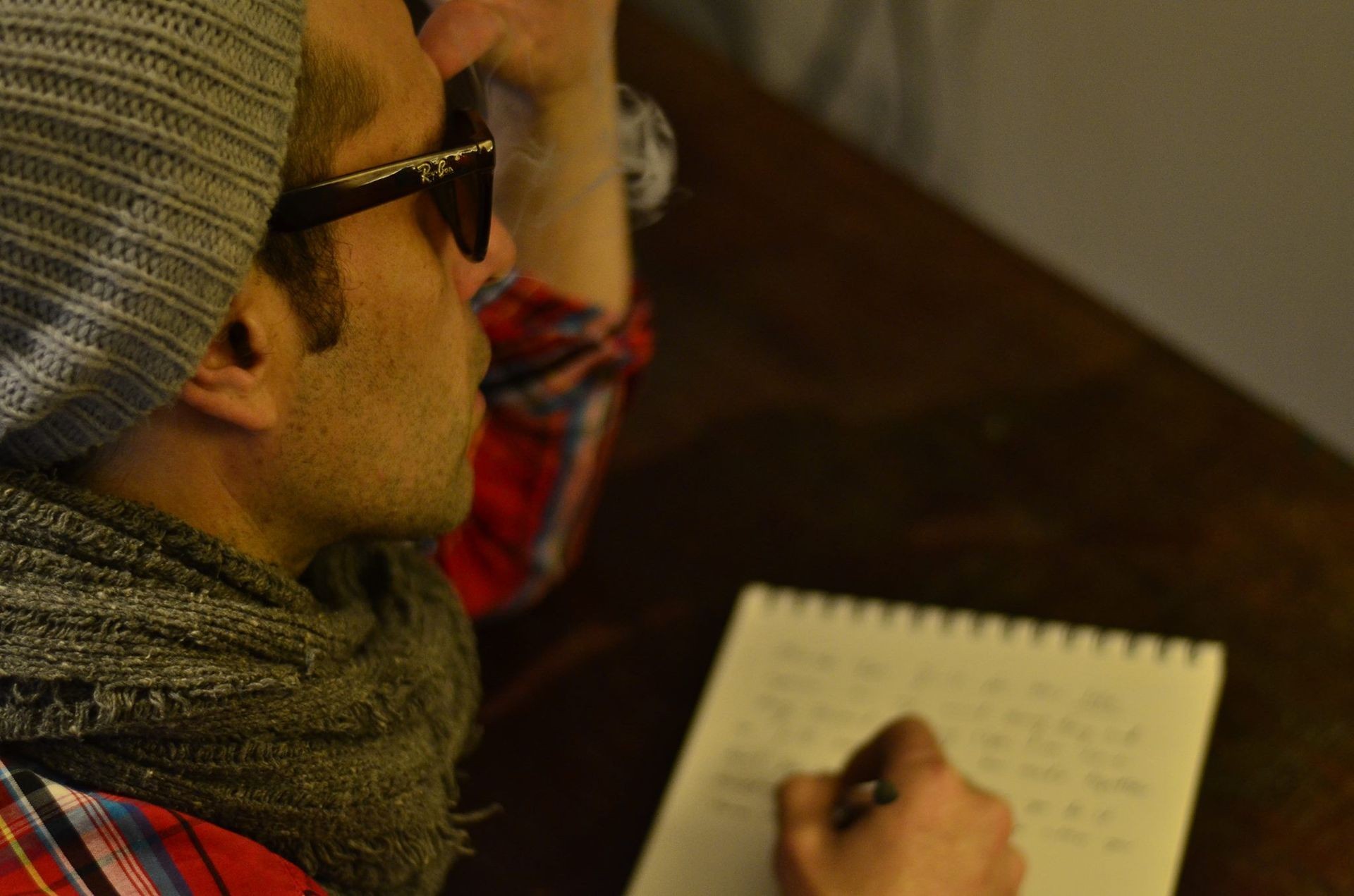 Person wearing a beanie and scarf, writing in a notebook on a wooden table.