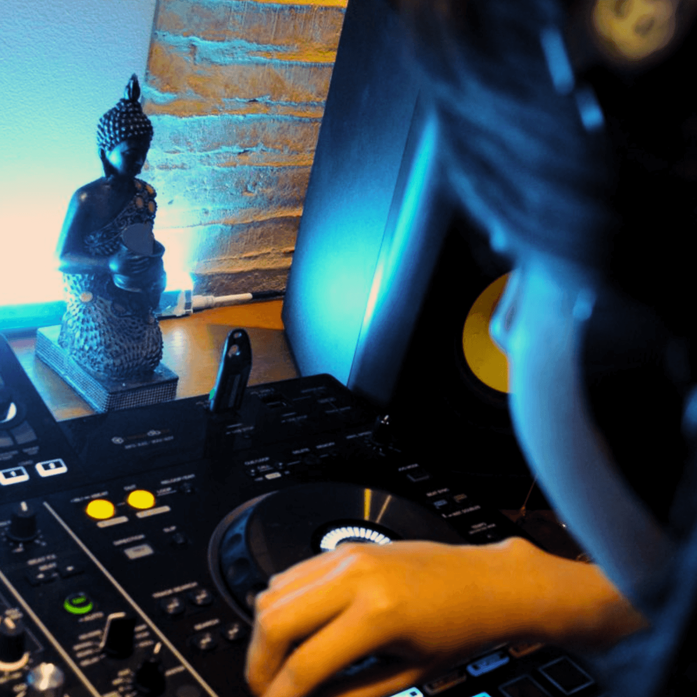 Person using DJ equipment with a Buddha statue and speaker in the background, illuminated by blue light.