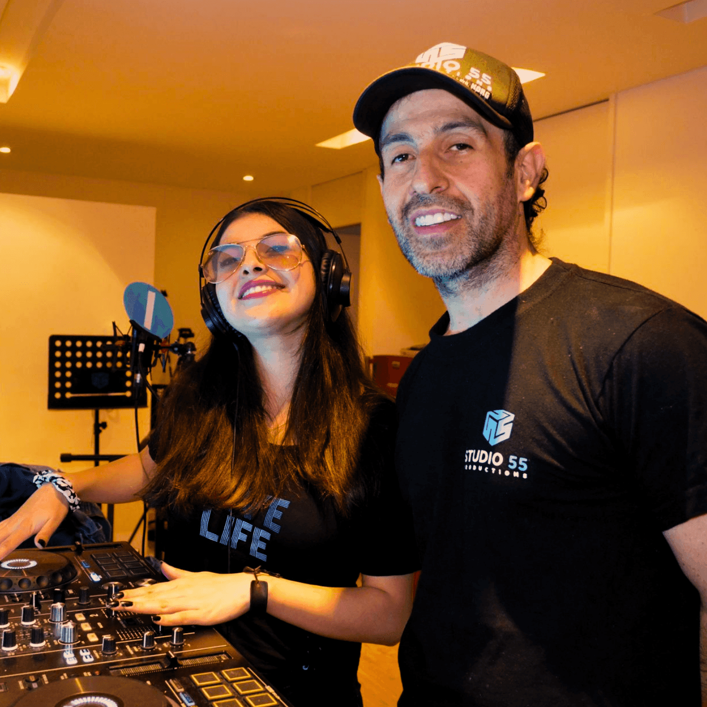 Two people smiling in a music studio, one using DJ equipment and the other standing beside.